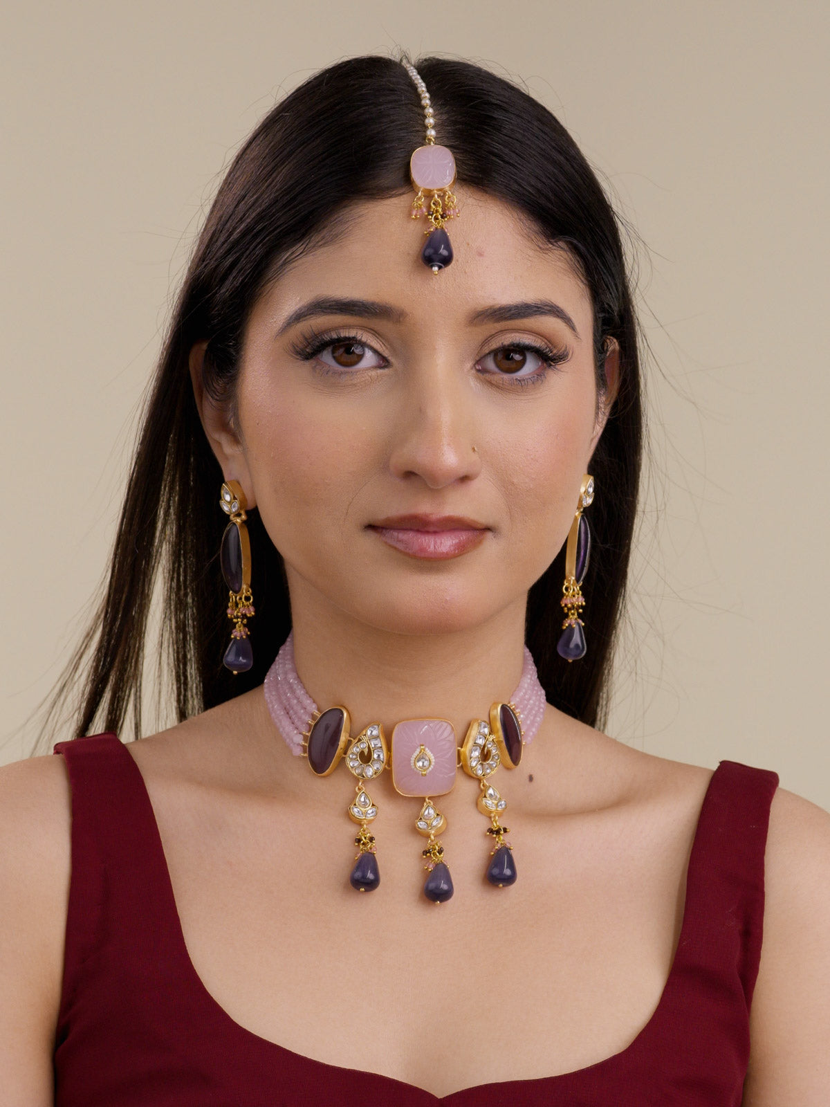 Model wearing the Pink Purple variant of a Fusion necklace set with matte gold tone detailing, pale pink bead collar strands, pale pink carved square centre, smoky purple side stones, clear stone accents, dark purple hanging drops, matching long earrings, and maang tikka.