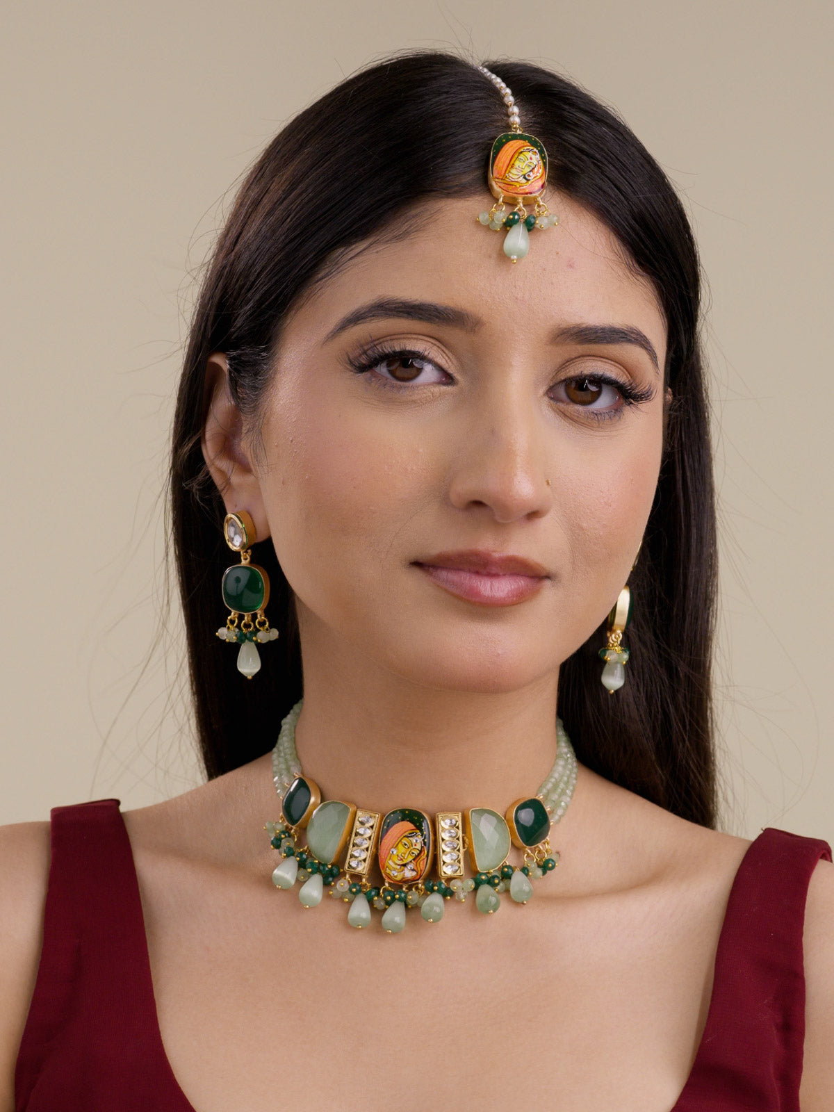 Model wearing a Fusion necklace set with matching drop earrings and maang tikka, sage green bead strands, sage green teardrop beads, dark green side stones, hand-painted centre face motif, kundan stone panels, and matte gold tone finish.