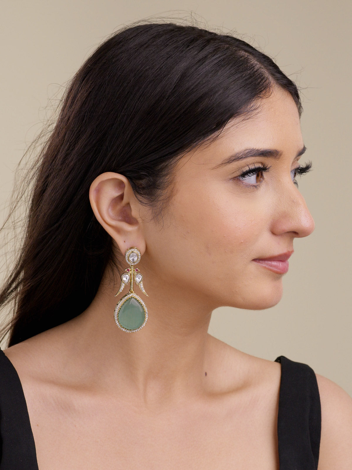 Model wearing a light antique gold tone Kundan earring with AD detailing, clear oval and teardrop stones, small ruby red accents, peacock-inspired side bird motifs, and a mint green teardrop lower stone in a side portrait shot.