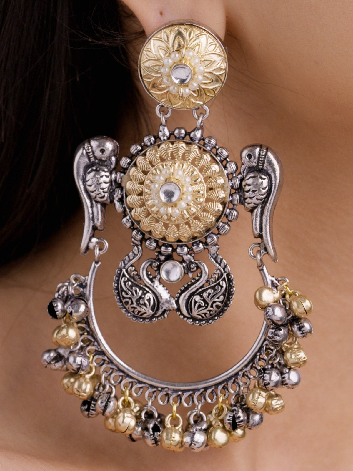 Close-up detail of statement oxidised multi tone earrings worn on a model ear, showing parrot-inspired side motifs, round gold-tone floral centres, pearl-style accents, and lower silver and gold bell drops.