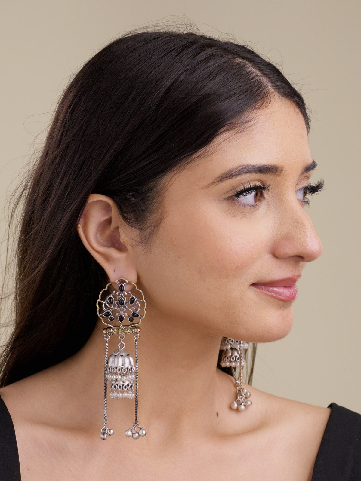 Model side profile wearing statement multi tone oxidised earrings with scalloped gold-tone openwork, black-colour stone details, silver jhumka bell drop, pearl bead accents, and long chain bell drops.