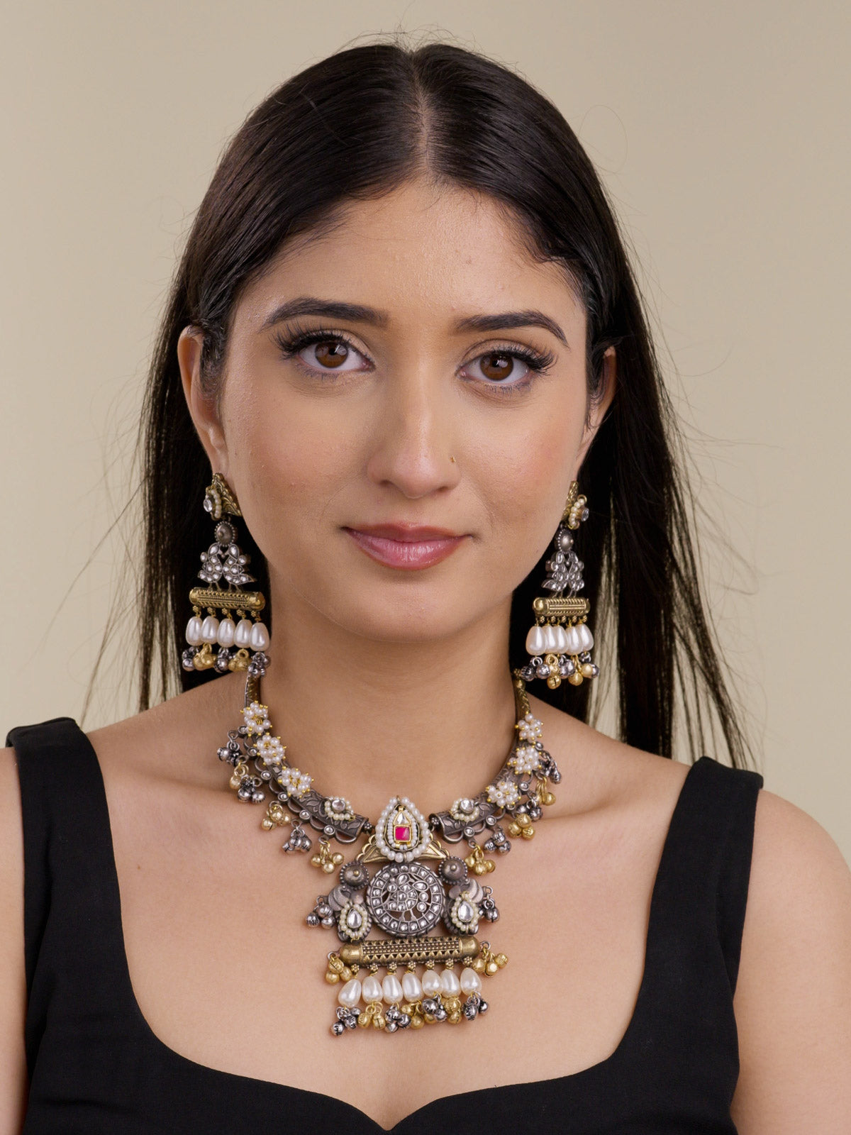 Model wearing a multi tone oxidised necklace set with a round centrepiece, Ruby Red accent stone, pearl drops, gold and oxidised bell drops, floral pearl clusters, and matching earrings in a front portrait shot.