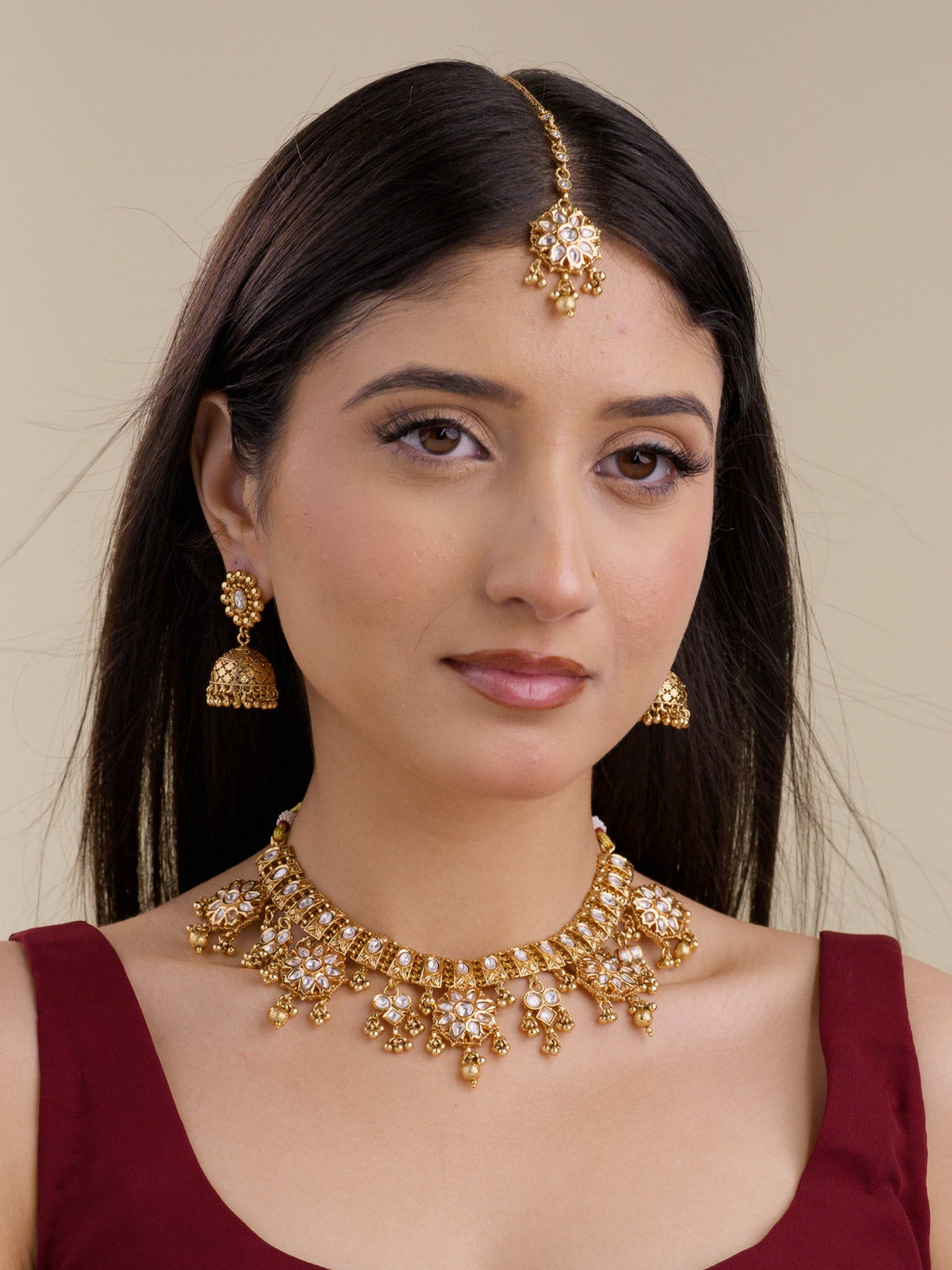 Model wearing a gold tone Rajwadi necklace set with kundan-look stones, floral drops, gold tone drop detailing, matching jhumka earrings, and a maang tikka.