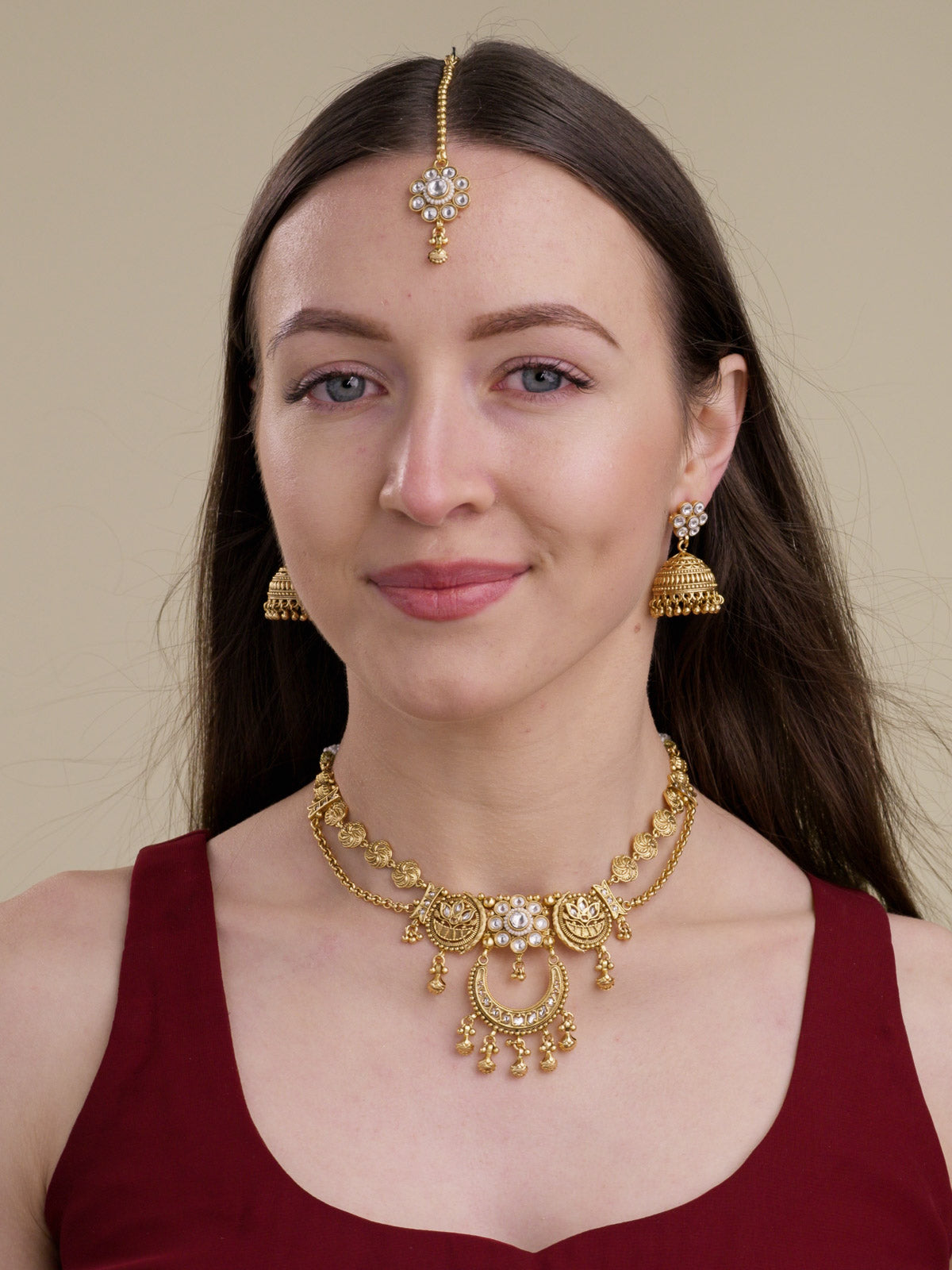 Matte gold tone Rajwadi necklace set with matching jhumka earrings and maang tikka, clear stone cluster details, crescent lower pendant, round medallion links, and small dangling metal drops on model front view.