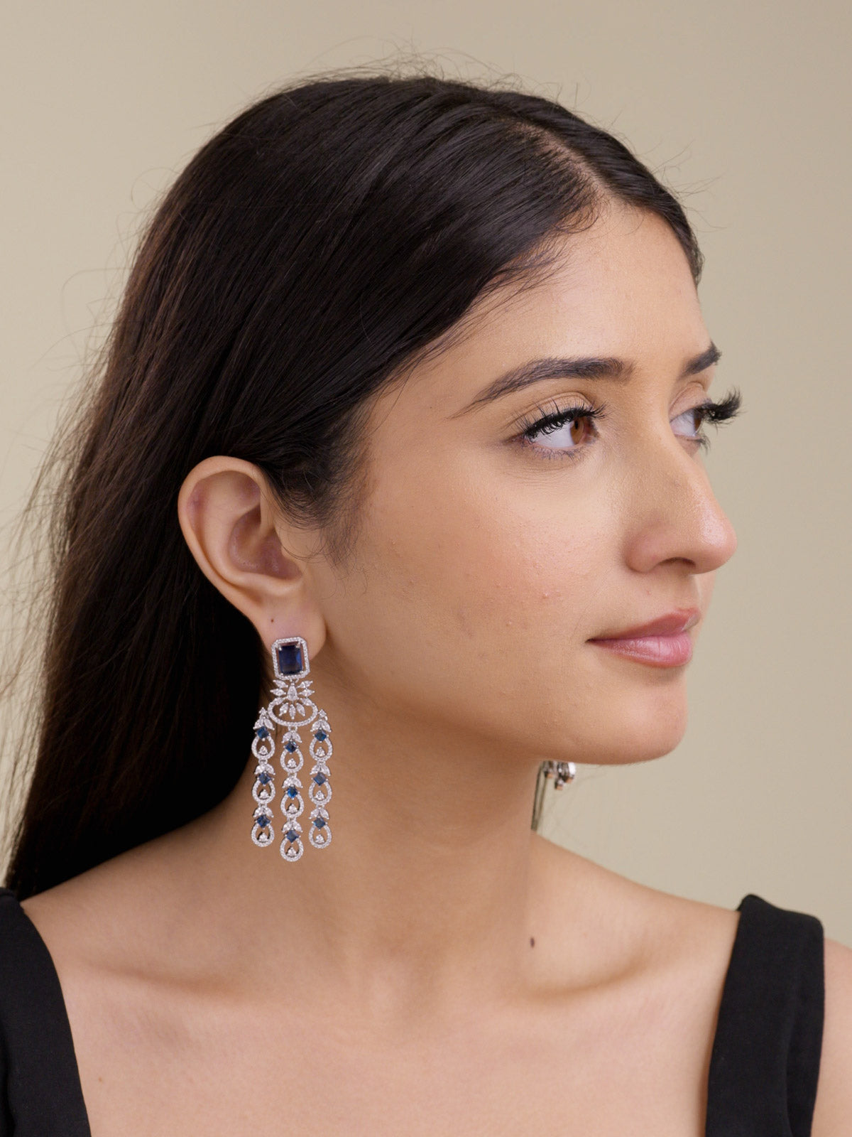 Model wearing a silver tone American Diamond earring with a blue square stud, clear CZ stones, curved centre accent, and three hanging teardrop strands with blue stone details in a side portrait shot.