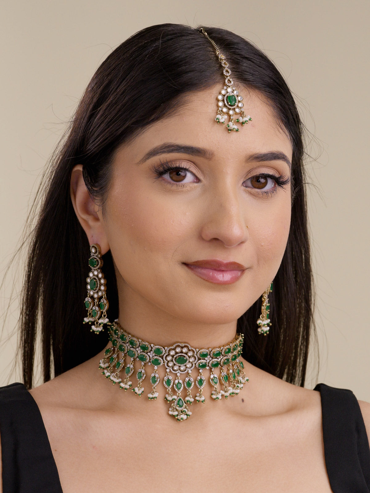 Model wearing an antique gold tone American Diamond choker necklace set with green stones, matching earrings, maang tikka, and pearl-style drops.