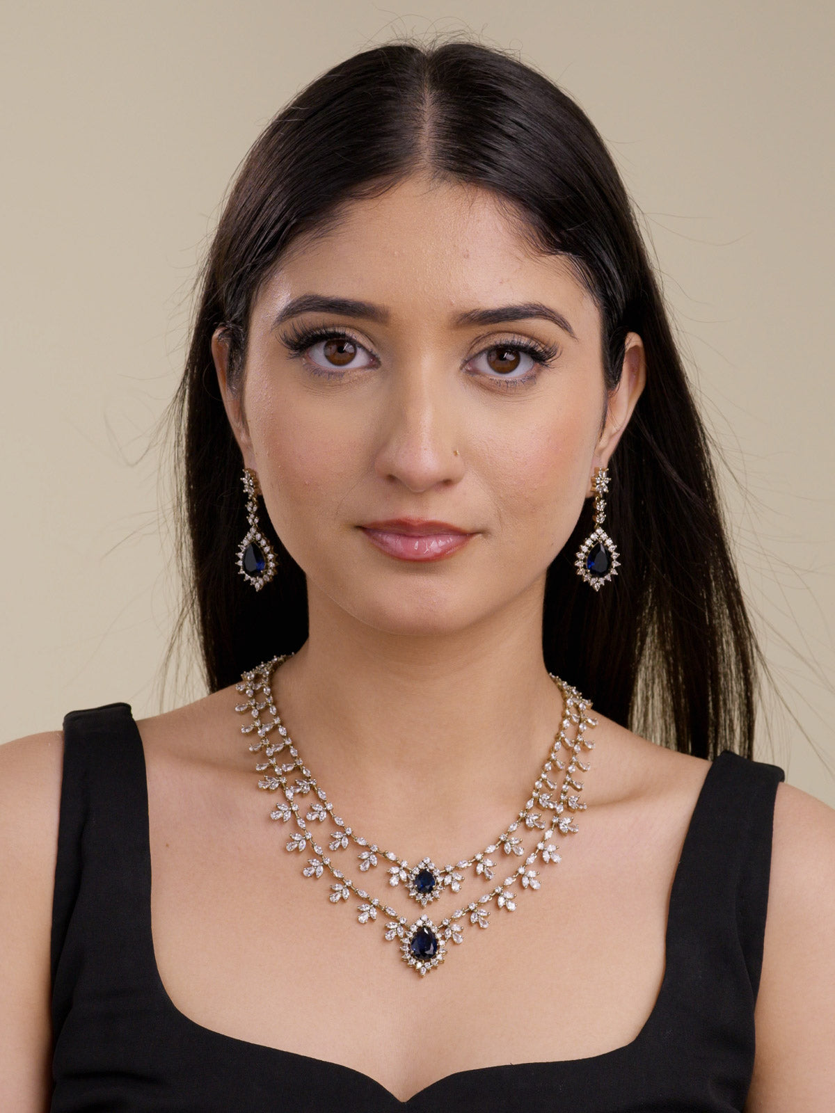 Model wearing an antique gold tone American Diamond necklace set with two-strand layered CZ stone trails, floral-style clusters, blue teardrop centre stones, and matching drop earrings in a front portrait shot.