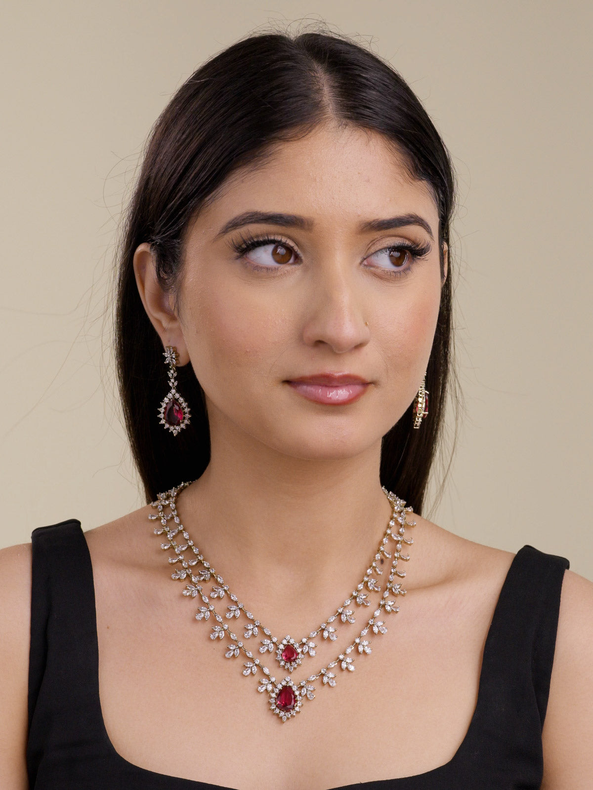 Model wearing an antique gold tone American Diamond necklace set with two-strand layered CZ stone trails, floral-style clusters, Ruby Red teardrop centre stones, and matching drop earrings in a three-quarter portrait shot.