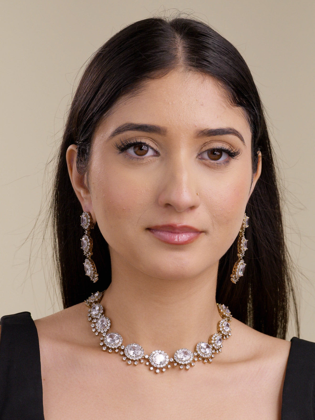 Front portrait of a model wearing a neutral American Diamond collar necklace set in antique gold tone with clear oval CZ centre stones, clear CZ halo detailing, fixed lower CZ accents, and matching long earrings.