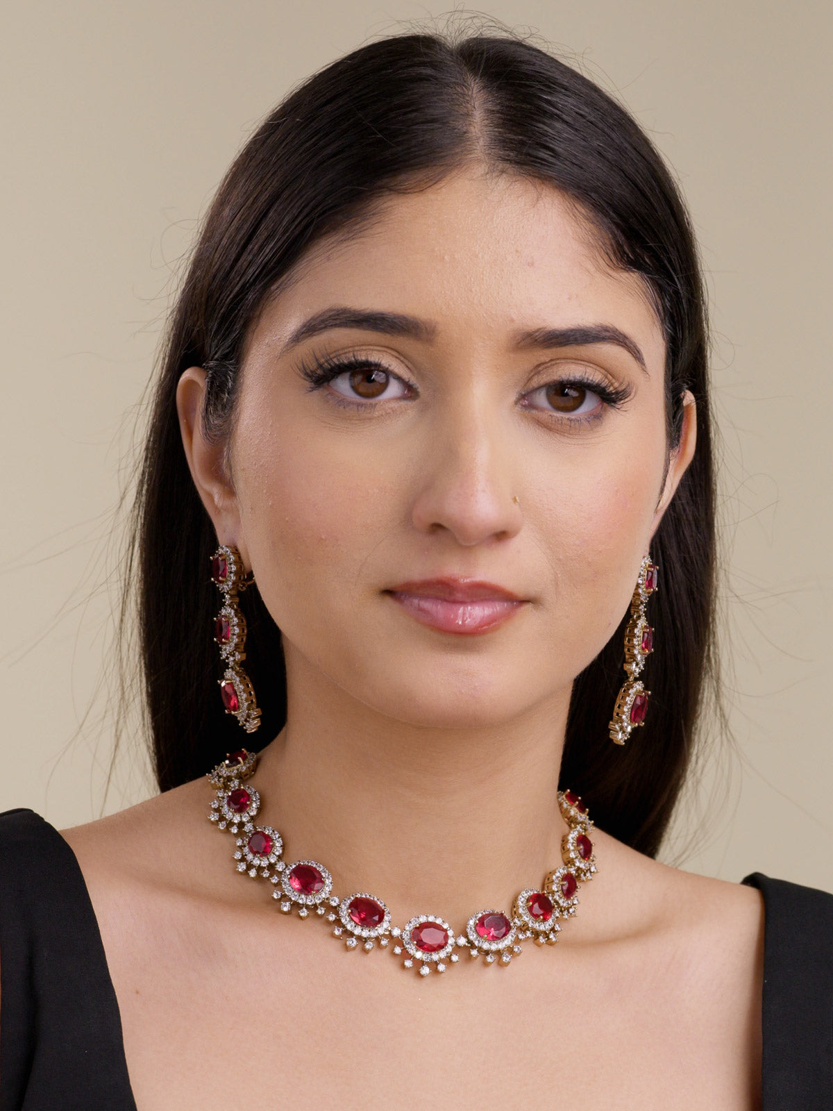 Front portrait of a model wearing a ruby red American Diamond collar necklace set in antique gold tone with oval CZ centre stones, clear CZ halo detailing, fixed lower CZ accents, and matching long earrings.