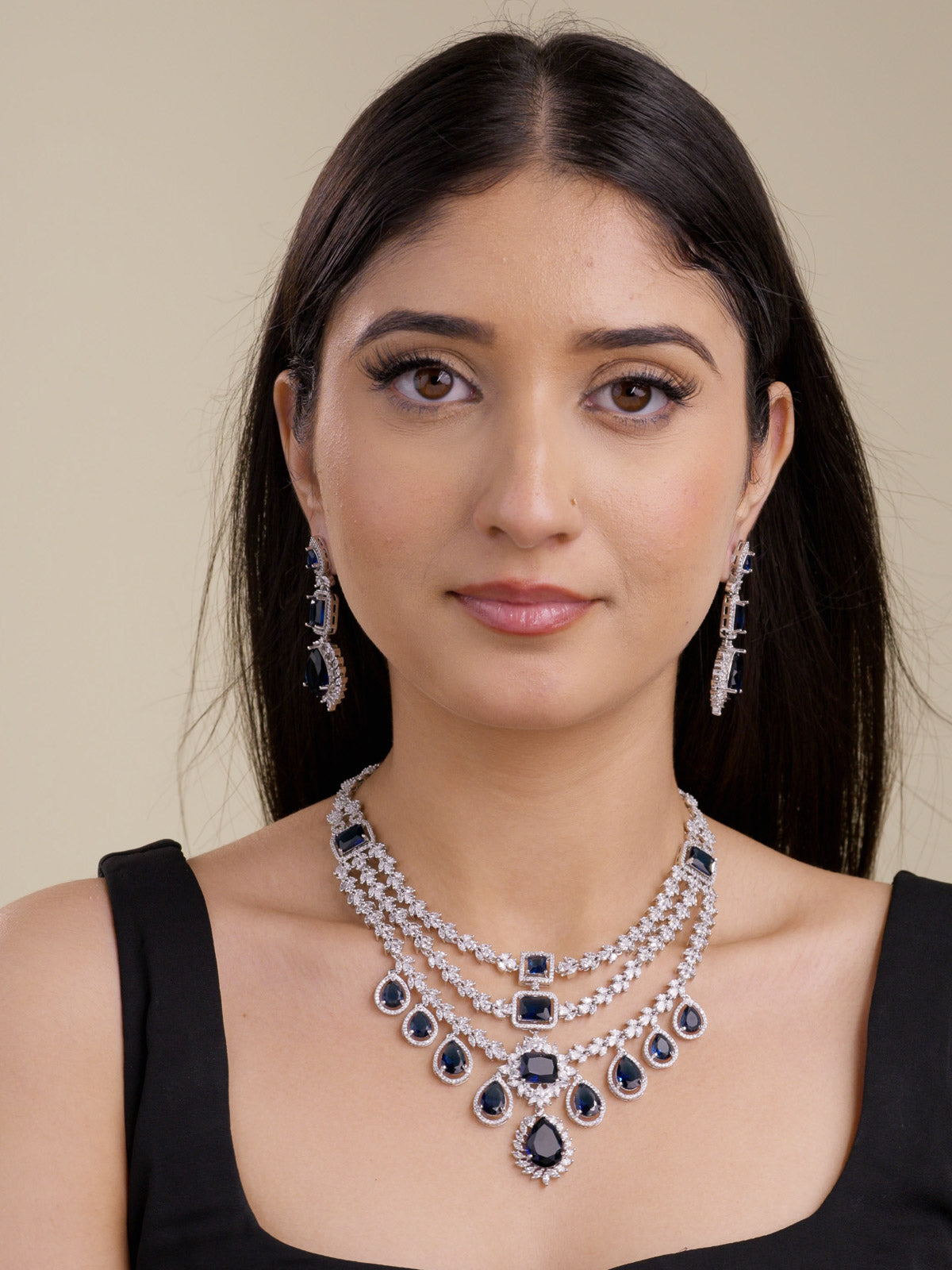 Front portrait of a model wearing a silver tone American Diamond necklace set with matching earrings, layered CZ stone detailing, deep blue rectangular and teardrop stones, and a centre drop pendant.
