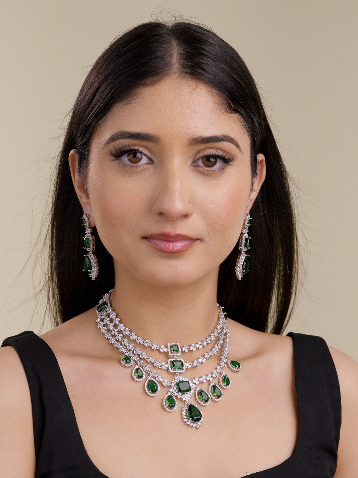 Front portrait of a model wearing a silver tone American Diamond necklace set with matching earrings, layered CZ stone detailing, green rectangular and teardrop stones, and a centre drop pendant.