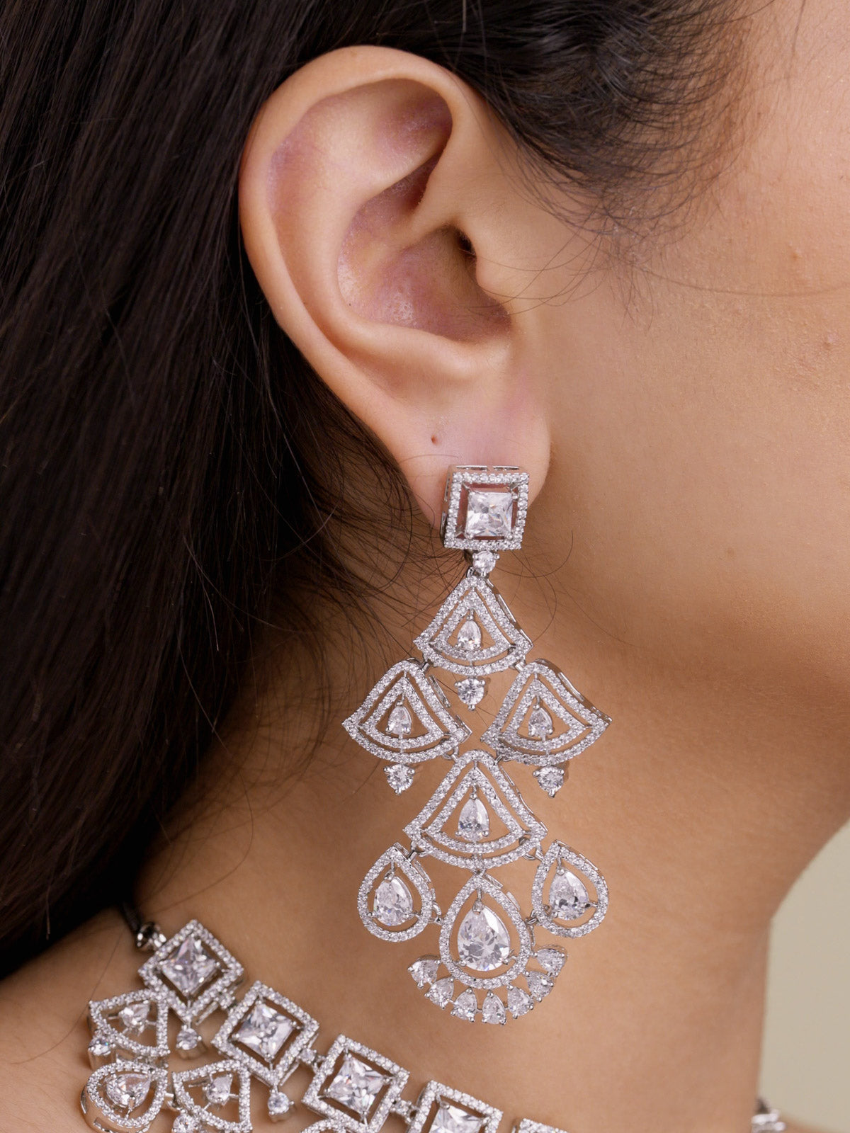Close-up side view of a model wearing a silver tone American Diamond long earring with a square CZ stud top, layered triangular CZ detailing, pear-shaped drops, and part of the matching choker-style necklace partially visible below.