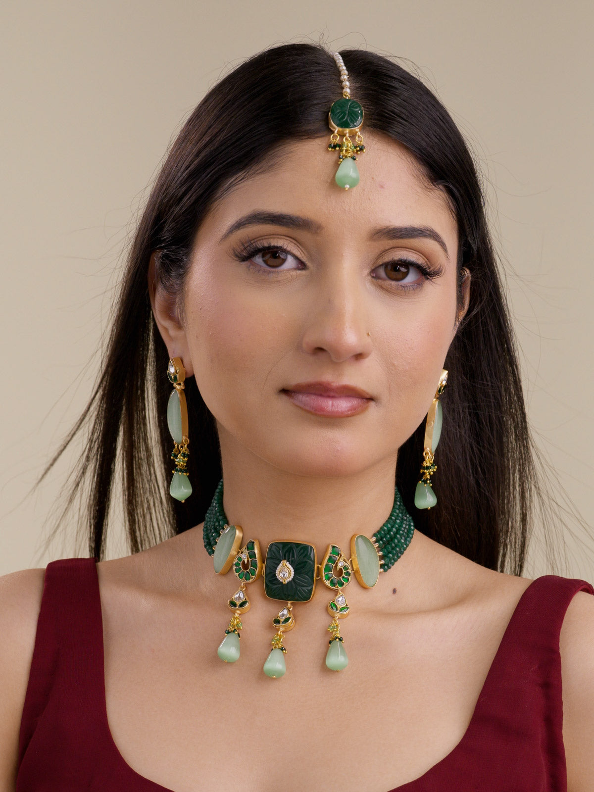 Model wearing the Green Sage variant of a Fusion necklace set with matte gold tone panels, dark green bead collar strands, carved dark green square centre, green paisley accents, sage green oval stones, sage green hanging drops, matching long earrings, and maang tikka.