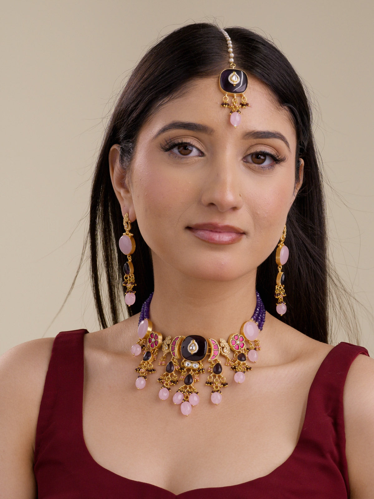 Front view of model wearing a Fusion necklace set with matching earrings and maang tikka, purple bead strands, matte gold tone detailing, pink oval accents, dark purple centre pieces, peacock motif studs, and hanging bead drops.