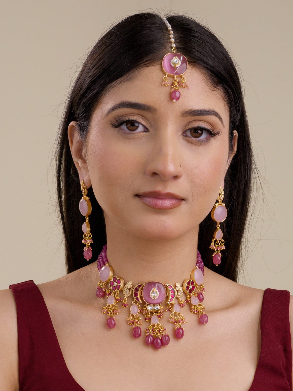 Front view of model wearing the Ruby Pink Fusion necklace set with matching earrings and maang tikka, pink bead strands, matte gold tone detailing, pale pink and ruby pink accents, peacock motif details, magenta floral sections, and hanging bead drops. 