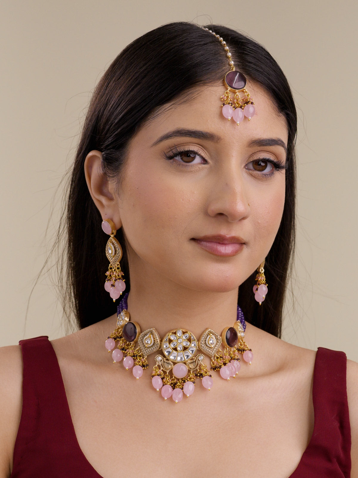 Front model view wearing Fusion necklace set in matte gold tone with white-stone floral centrepiece, dark purple round stones, lattice teardrop motifs, purple bead strands, pale pink bead drops, matching earrings, and maang tikka.