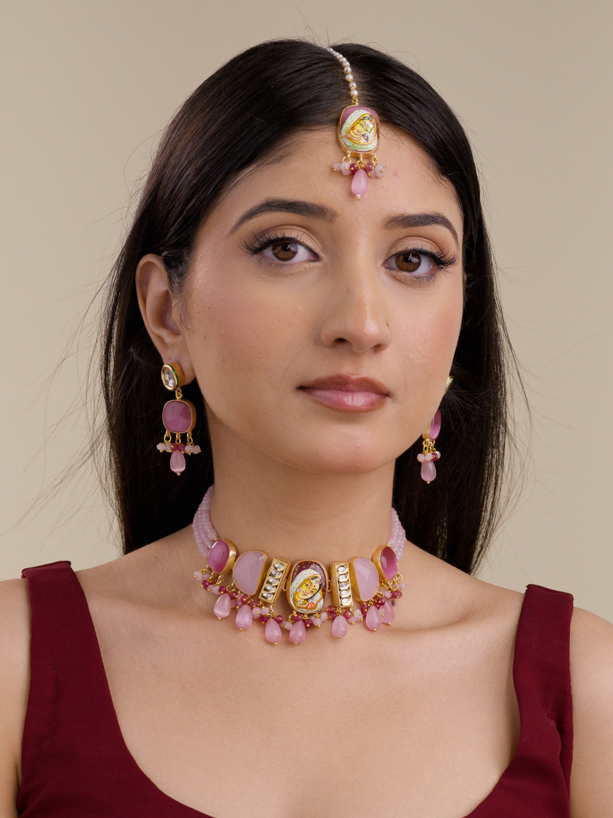 Model wearing a Fusion necklace set with matching earrings and maang tikka, pink and ruby bead drops, hand-painted centre face motif, kundan stone panels, and matte gold tone finish.