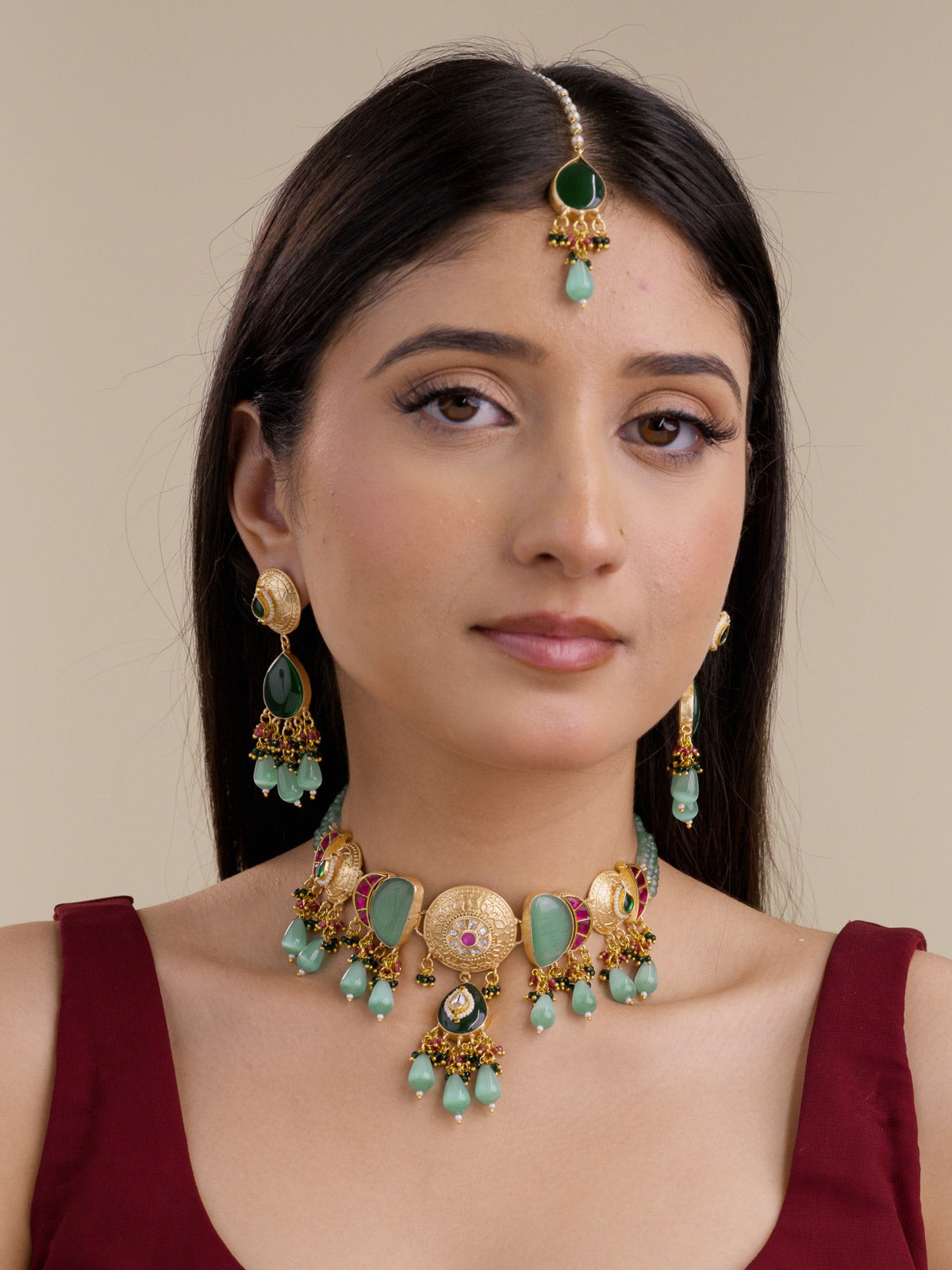 Model wearing the Mint Ruby Fusion necklace set in matte gold tone with matching earrings and maang tikka, showing round textured medallions, green stone panels, ruby bead accents, mint teardrop drops, and bead cluster detailing.