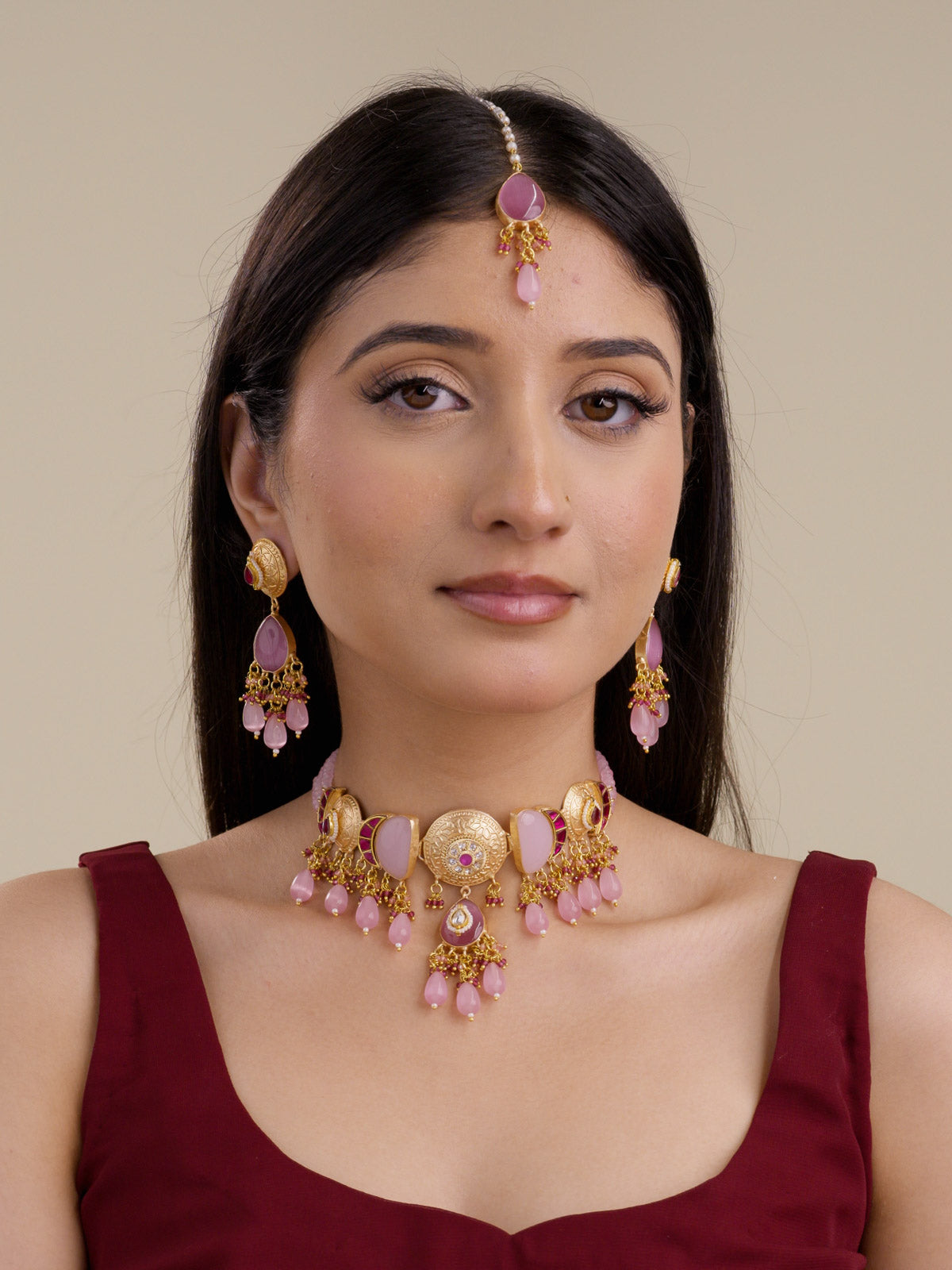 Model wearing the Pink Ruby Fusion necklace set in matte gold tone with matching earrings and maang tikka, showing round textured medallions, soft pink stone panels, ruby bead accents, pink teardrop drops, and bead cluster detailing.