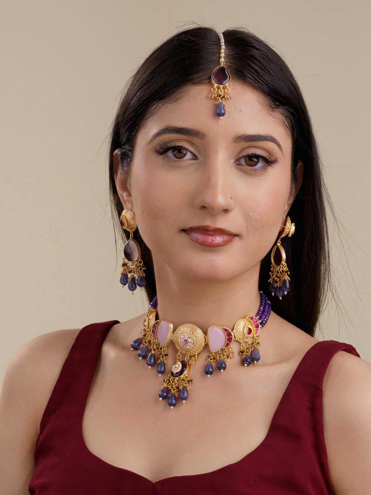 Model wearing the Purple Ruby Fusion necklace set in matte gold tone with matching earrings and maang tikka, showing round textured medallions, soft pink stone panels, ruby bead accents, purple teardrop drops, purple bead strands, and bead cluster detailing.