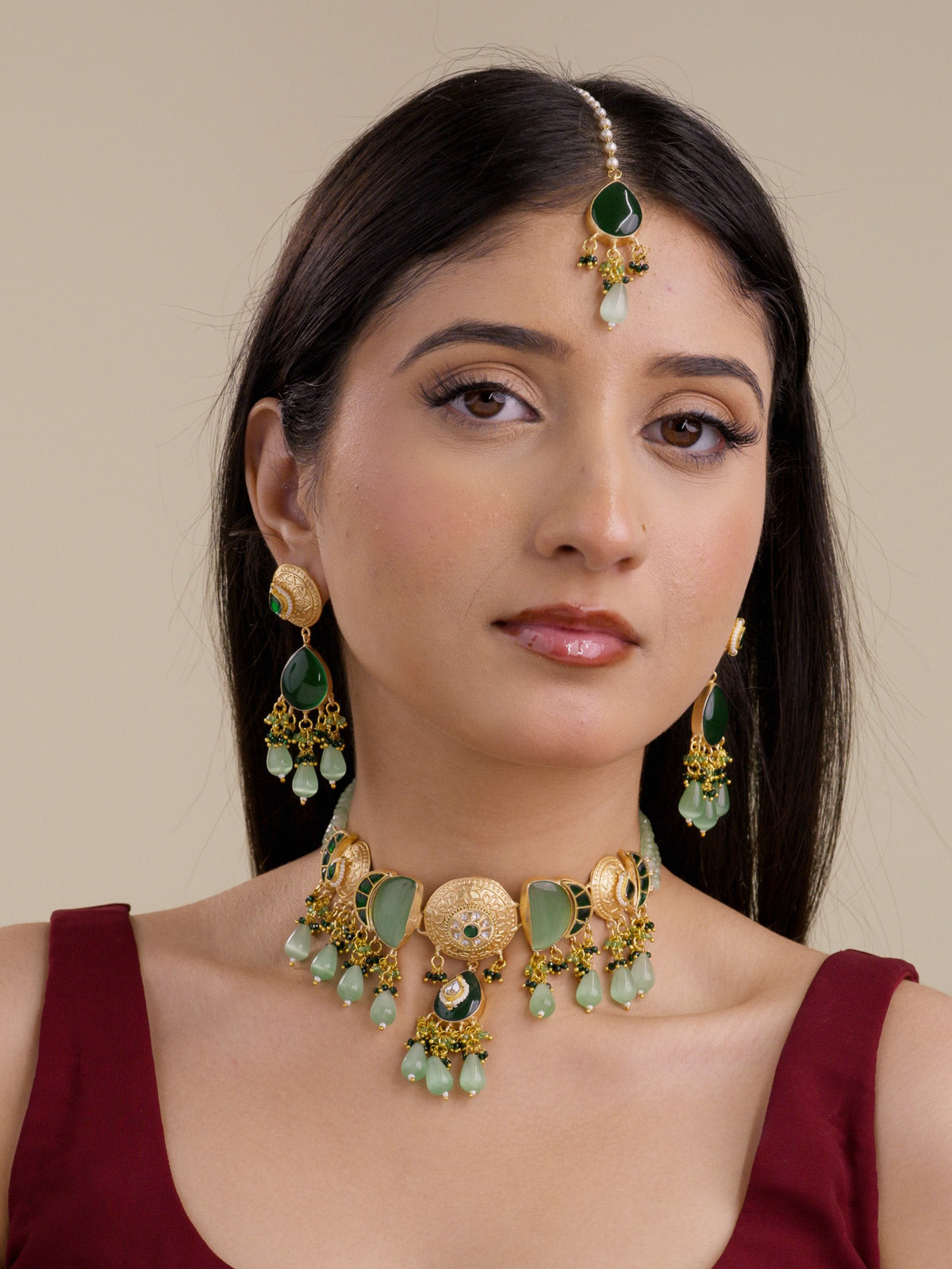Model wearing the Sage Green Fusion necklace set in matte gold tone with matching earrings and maang tikka, showing round textured medallions, green stone panels, sage green teardrop drops, dark green bead clusters, and layered sage bead strands.