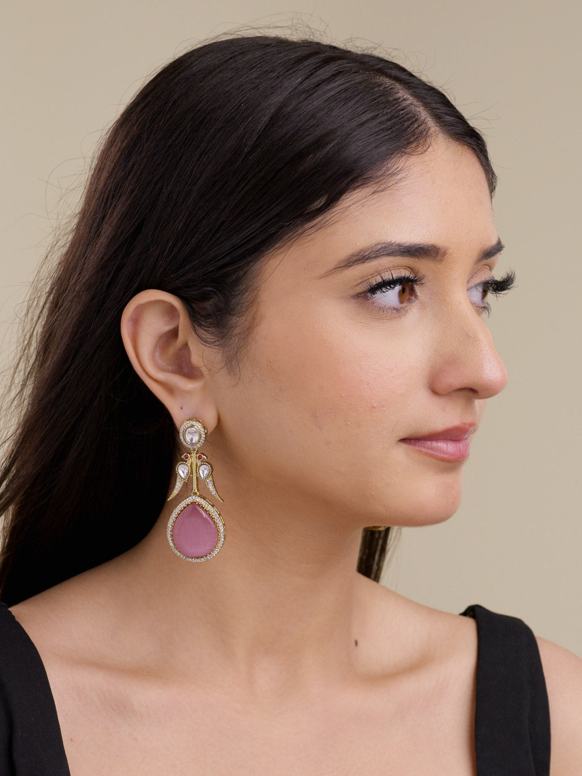 Model wearing a light antique gold tone Kundan earring with AD detailing, clear oval and teardrop stones, small ruby red accents, peacock-inspired side bird motifs, and a pink teardrop lower stone in a side portrait shot.