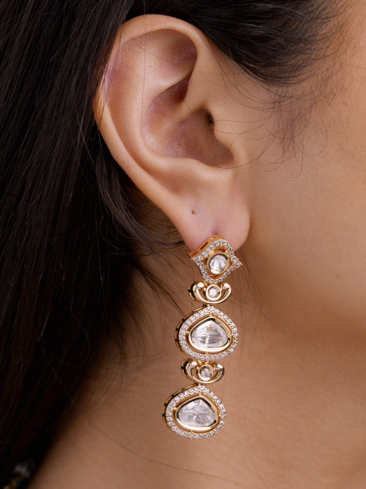 Close-up of matching Kundan drop earring in champagne gold tone on a model ear, showing angled stud top, two teardrop kundan-look stones, curved link accents, and American Diamond detailing.