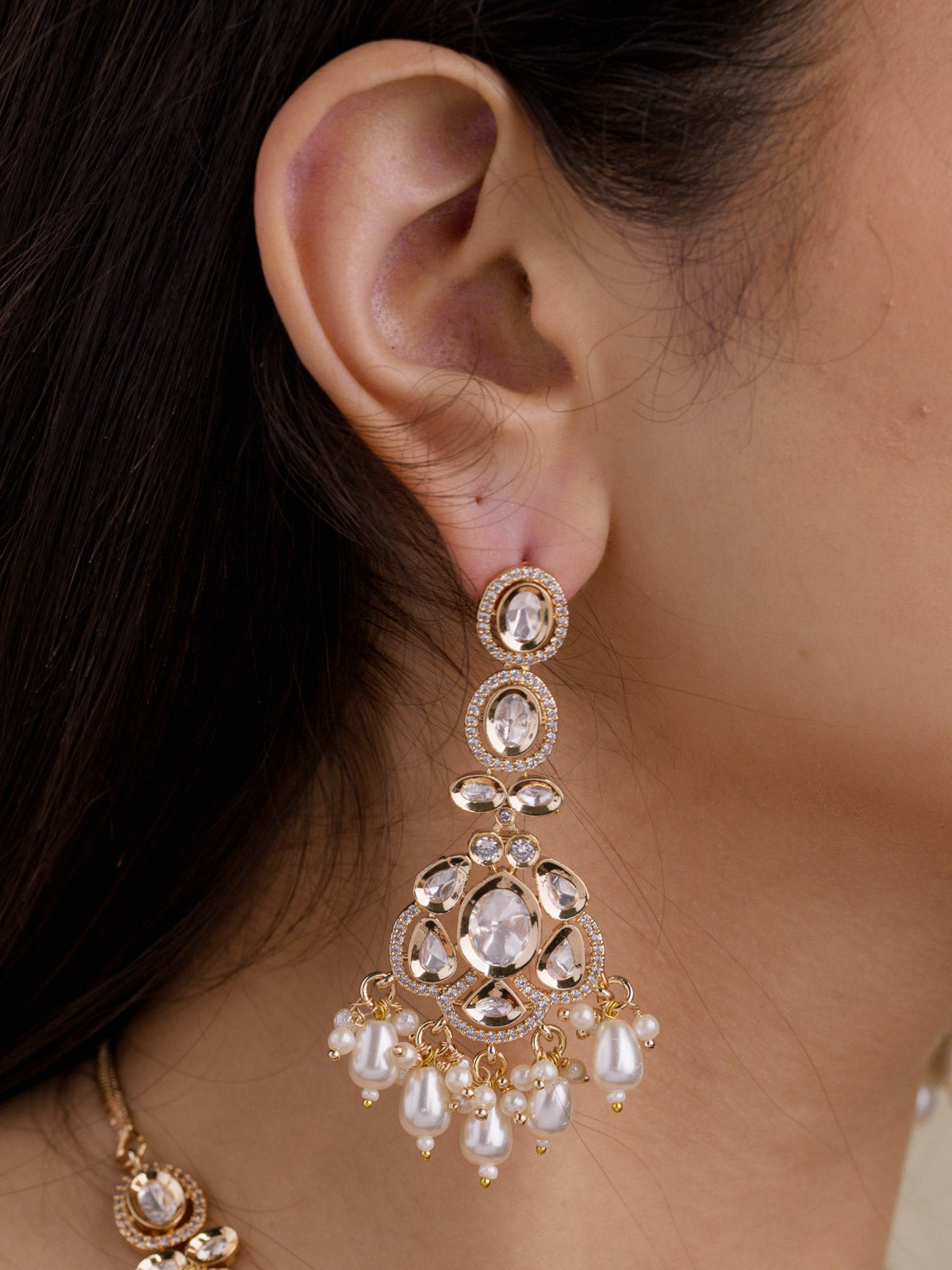 Close-up of matching champagne gold tone earring with oval kundan-look stones, American Diamond-style borders, teardrop lower detail, pearl-style drops, and partial necklace detail.