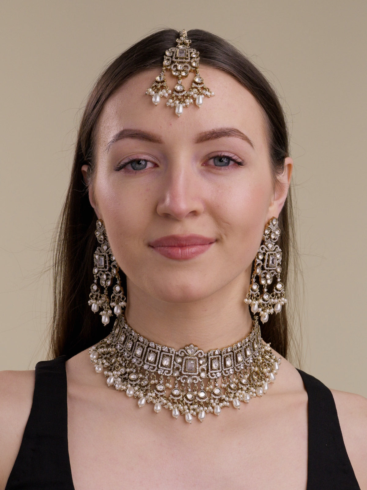Front view of a model wearing a neutral Kundan choker-style necklace set in Antique Gold Tone with matching long earrings, maang tikka, rectangular frame detailing, kundan-look centre stones, American Diamond accents, and pearl drops.