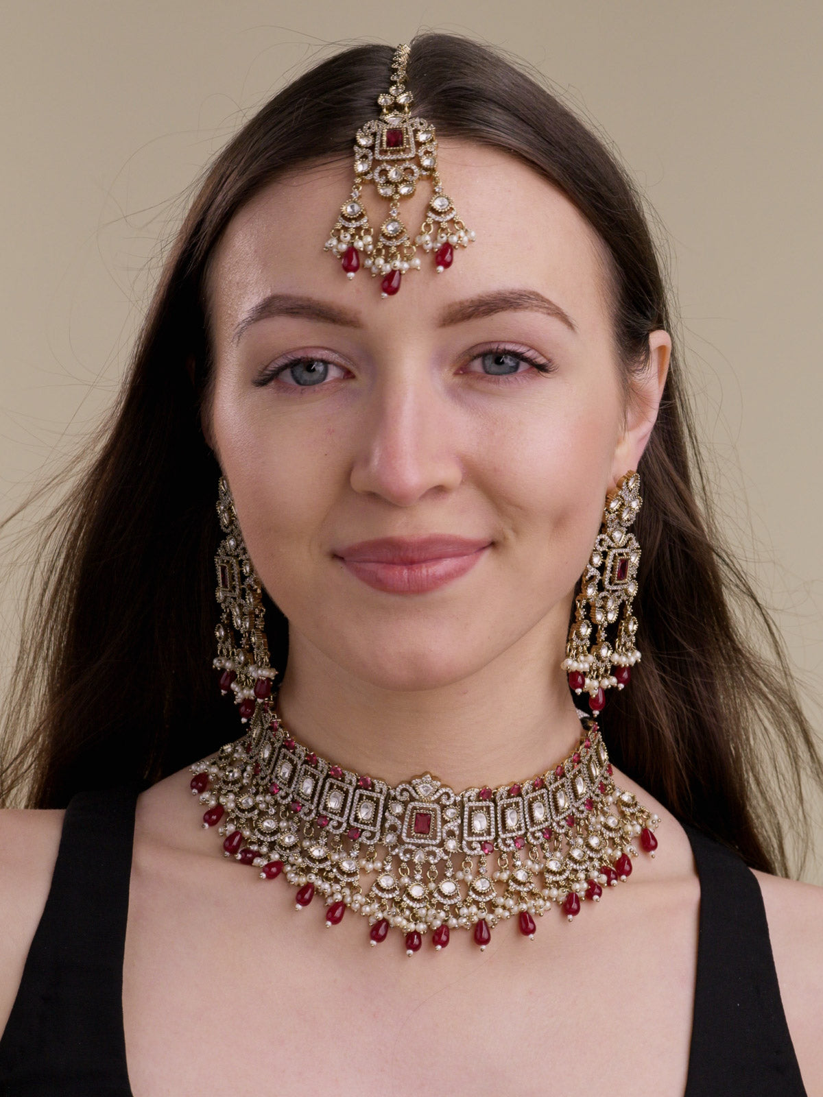 Front view of a model wearing a Ruby Red Kundan choker-style necklace set in Antique Gold Tone with matching long earrings, maang tikka, rectangular frame detailing, kundan-look centre stones, American Diamond accents, Ruby Red drops, and pearl accents.