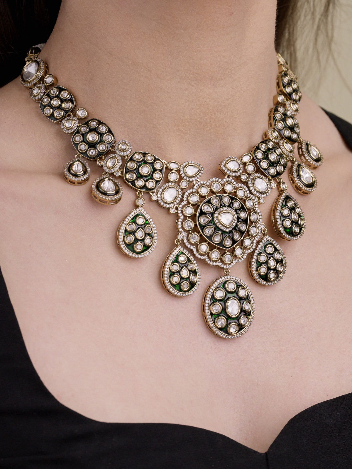 Close-up of a model wearing an antique gold tone Kundan necklace set with green enamel-look detailing, kundan-look stones, CZ accents, and matching earrings partly visible.