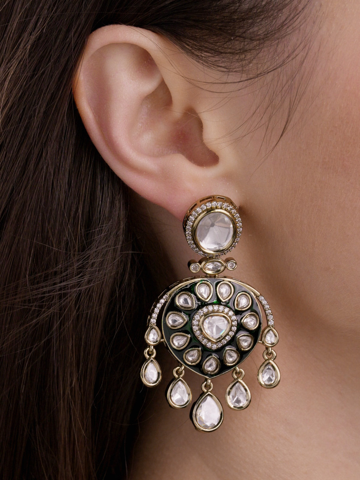 Close-up of a model wearing an antique gold tone Kundan earring with green enamel-look detailing, kundan-look stones, CZ accents, and hanging teardrop drops.