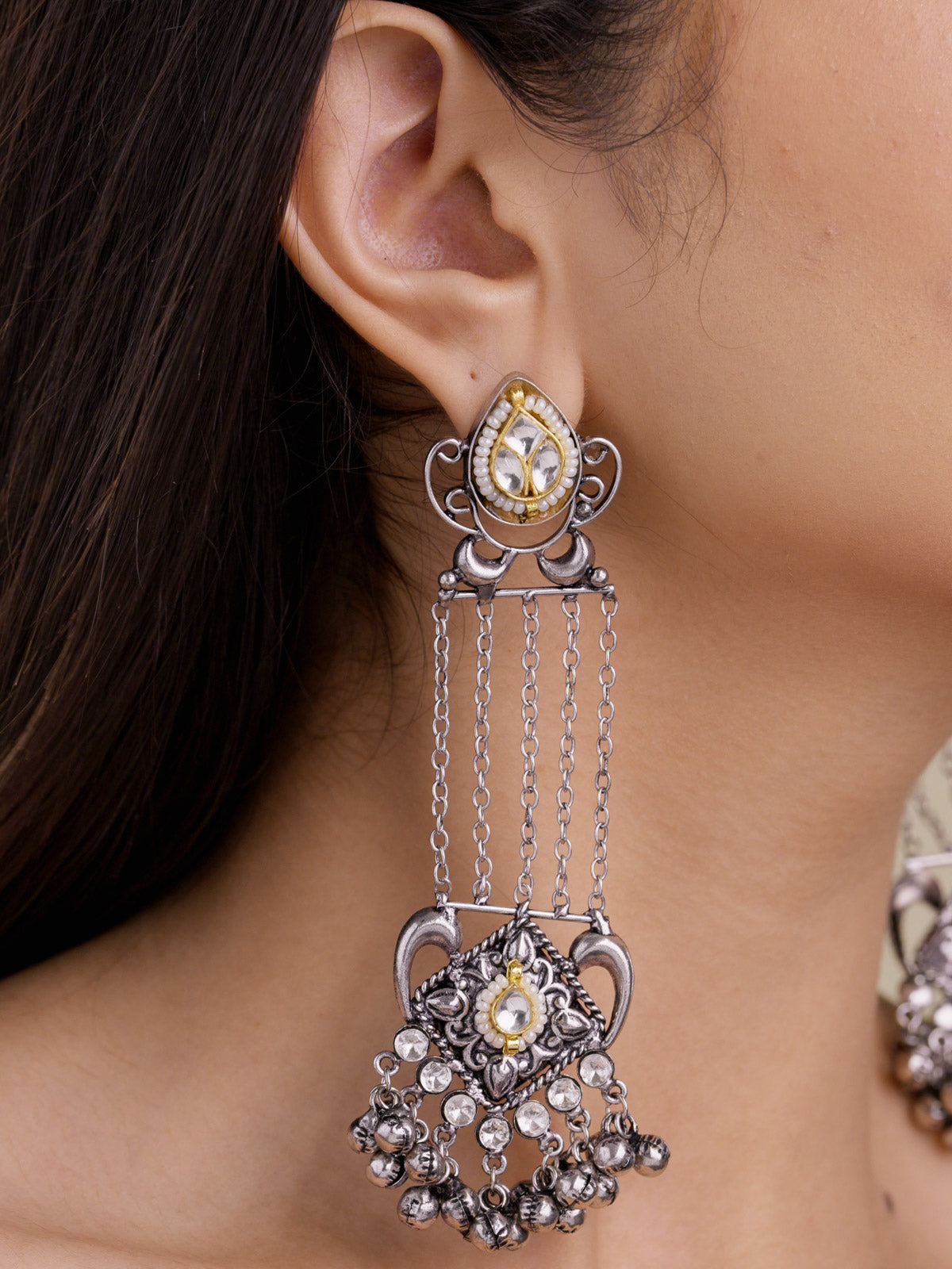 Close-up of model wearing a long oxidised earring with a teardrop kundan-look stud, open scroll detailing, hanging chain strands, carved lower panel, clear stone accents, and metal bell fringe.