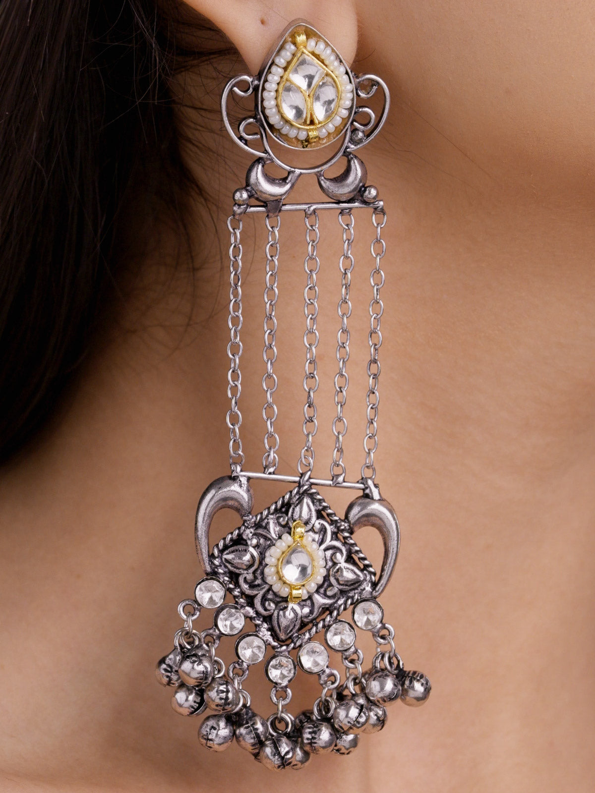 Close-up detail of a long oxidised earring with a teardrop kundan-look stud, pearl-look border, hanging chain strands, carved lower panel, clear stone accents, and metal bell fringe.