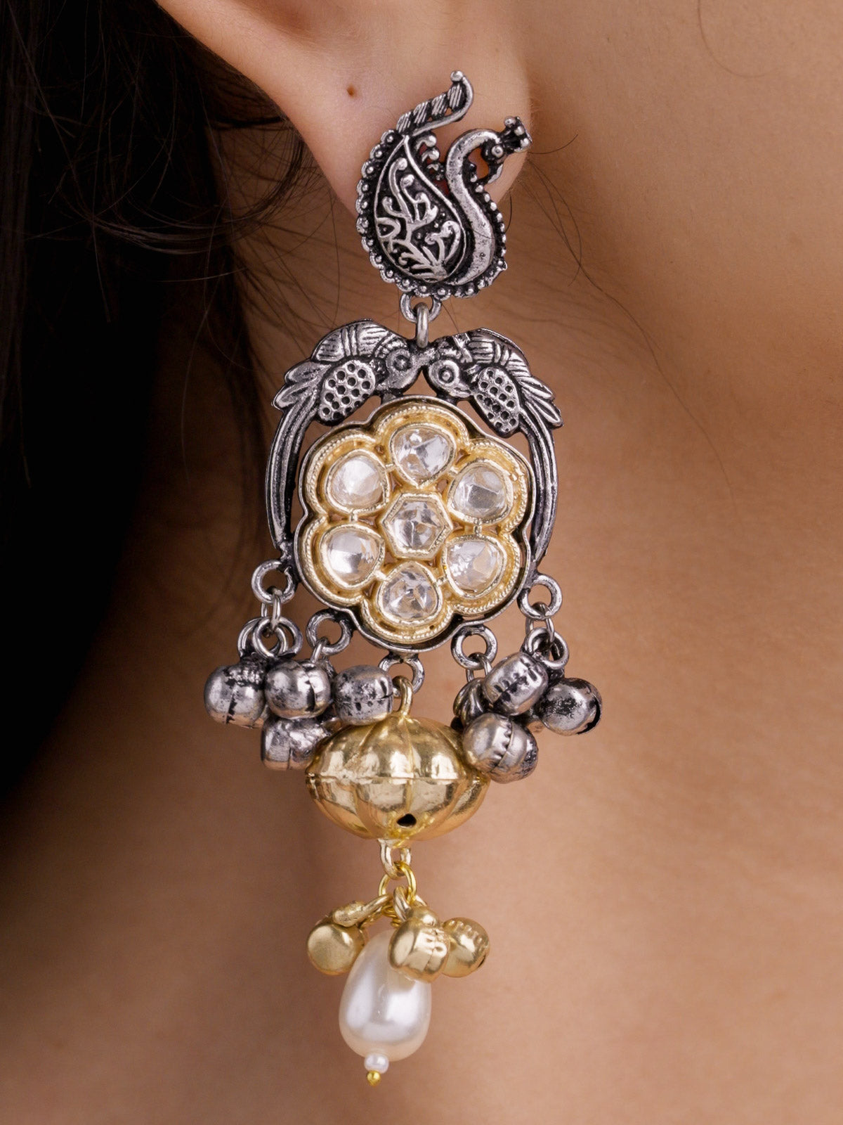 Close-up of an oxidised Multi Tone earring on a model ear with a peacock-inspired paisley stud motif, bird-like upper detailing, round gold-tone kundan-look stone centre, silver bead drops, gold-tone bell detail, and pearl-look lower drop.