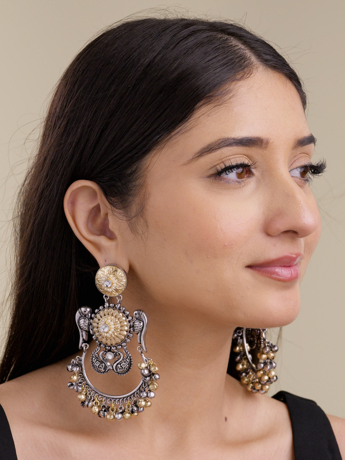 Model wearing statement oxidised multi tone earrings, with the front earring showing parrot-inspired side motifs, round gold-tone floral centres, pearl-style accents, and lower silver and gold bell drops.