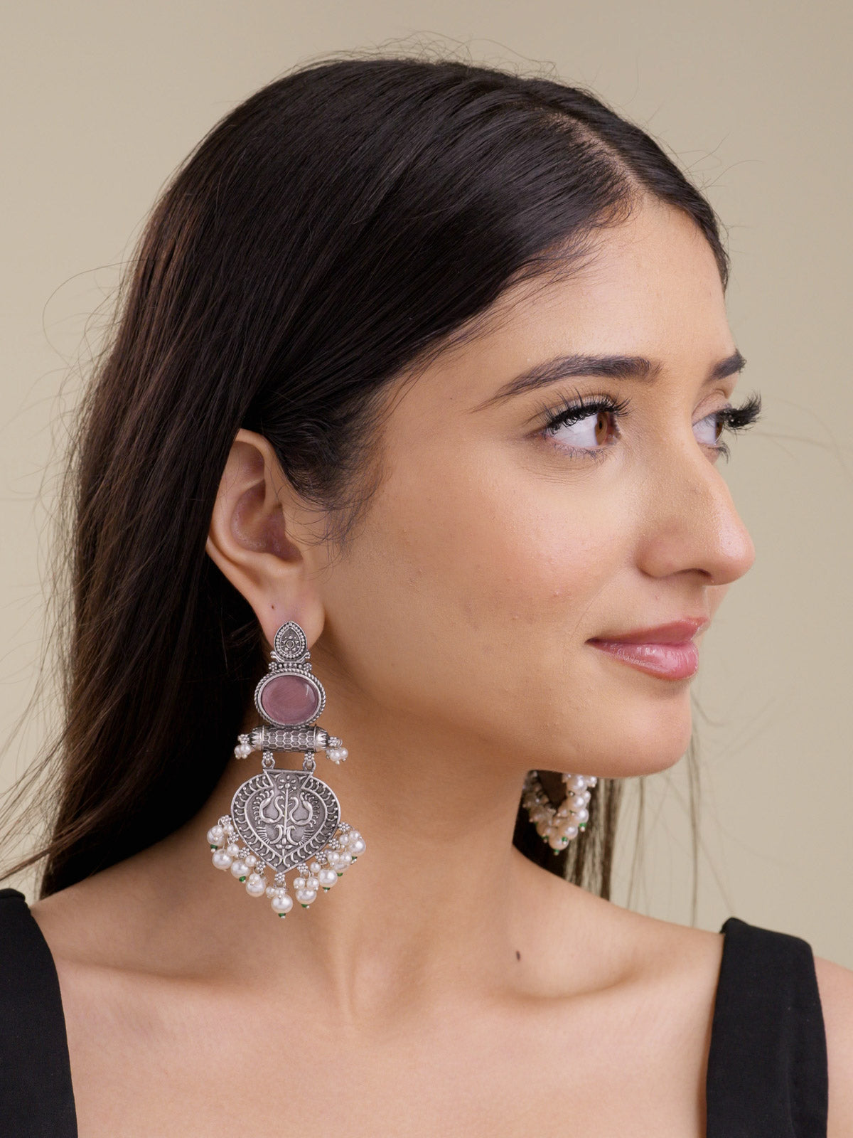 Model wearing statement oxidised earrings with upper pink oval stones, carved paan-leaf lower medallions, pearl-style hanging drops, and small green bead accents.