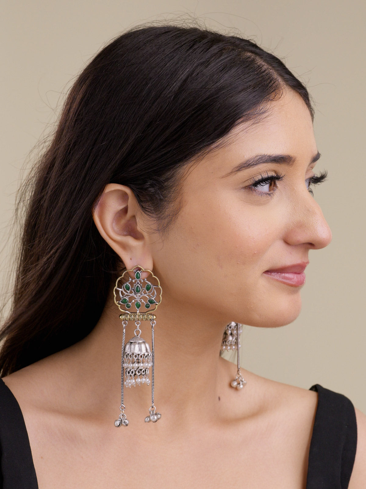 Model side profile wearing statement multi tone oxidised earrings with scalloped gold-tone openwork, green-colour stone details, silver jhumka bell drop, pearl bead accents, and long chain bell drops.