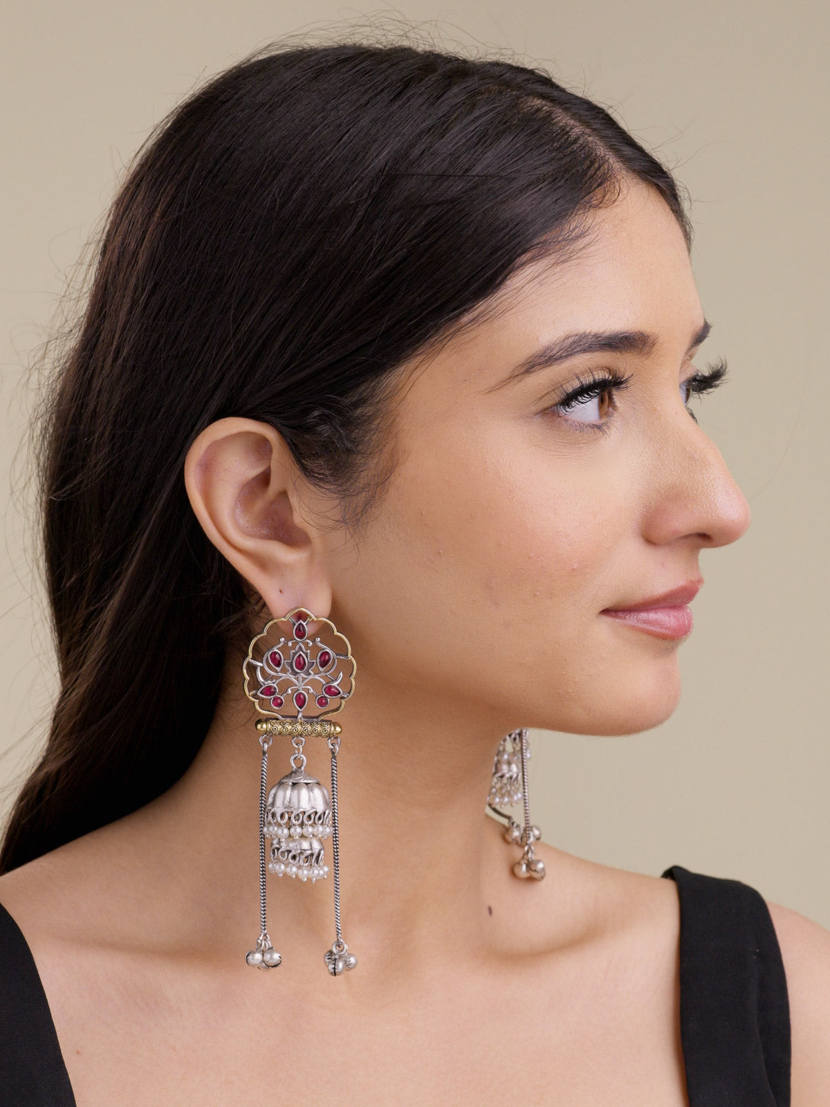 Model side profile wearing statement multi tone oxidised earrings with scalloped gold-tone openwork, ruby-colour stone details, silver jhumka bell drop, pearl bead accents, and long chain drops.
