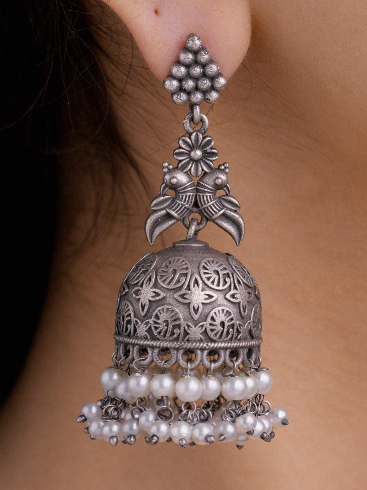 Close-up of a model wearing an oxidised jhumka earring with a clustered metal bead stud, floral and paired bird motif detail, carved dome, and pearl drops.