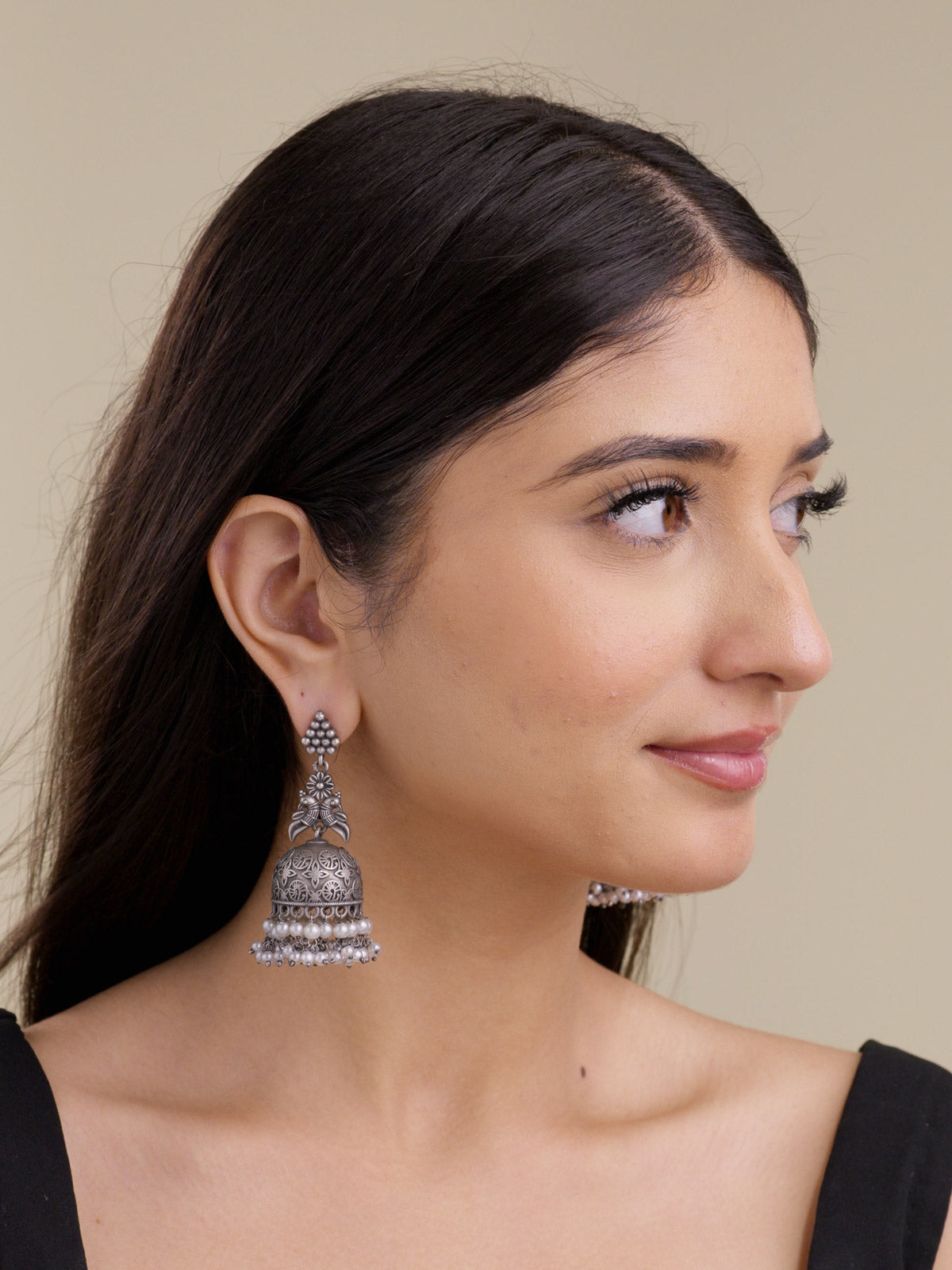 Model wearing an oxidised jhumka earring with a clustered metal bead stud, floral and paired bird motif detail, carved dome, and pearl drops in a portrait shot.
