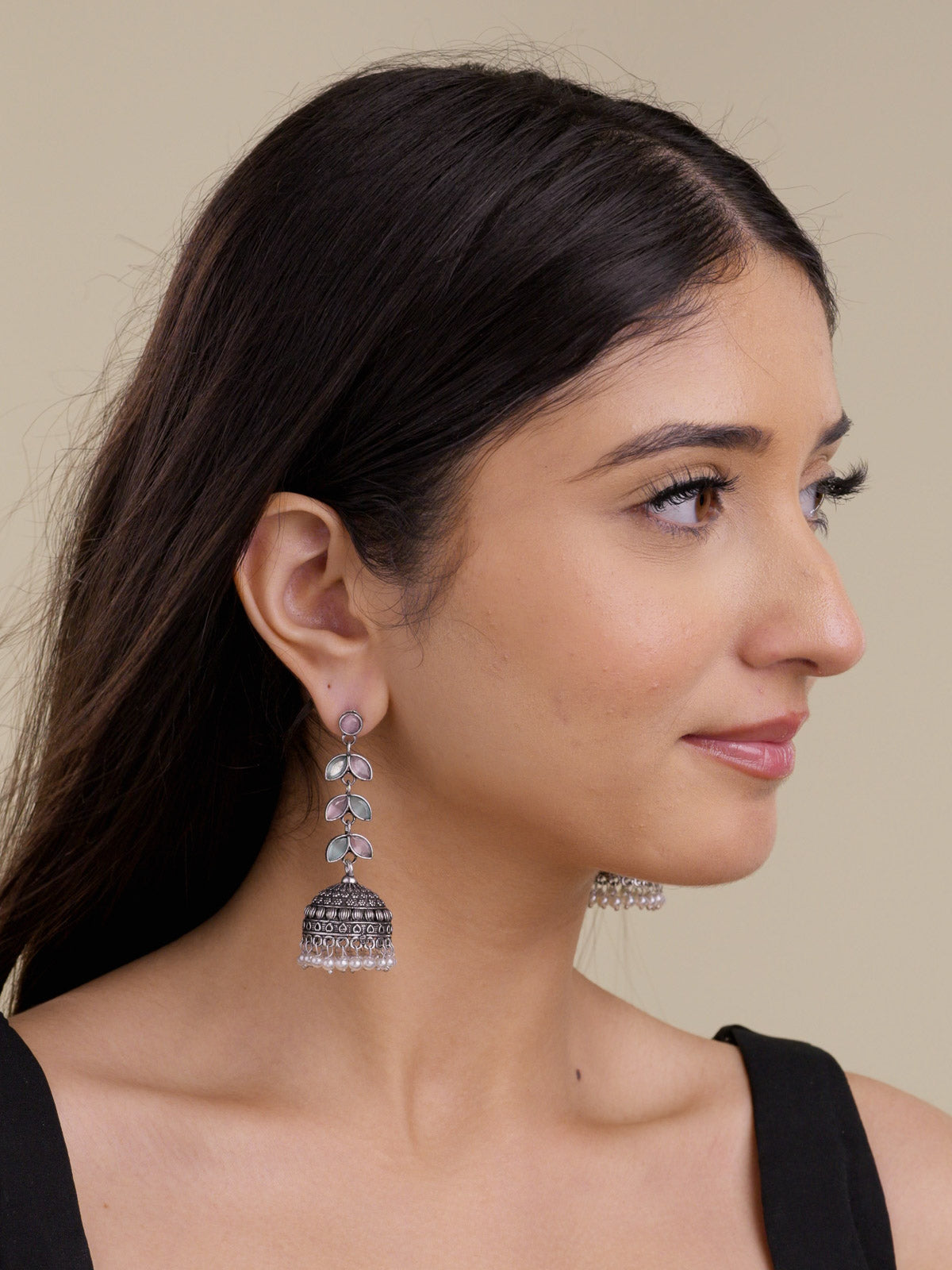Model wearing oxidised jhumka earrings with pink and mint petal-shaped stones, textured dome detail, and pearl drops in a portrait shot.