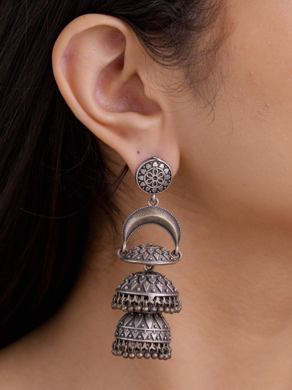 Close-up side view of a model wearing an oxidised tiered jhumka earring with a round floral stud, crescent arch connector, patterned double jhumka domes, and small round metal drops.