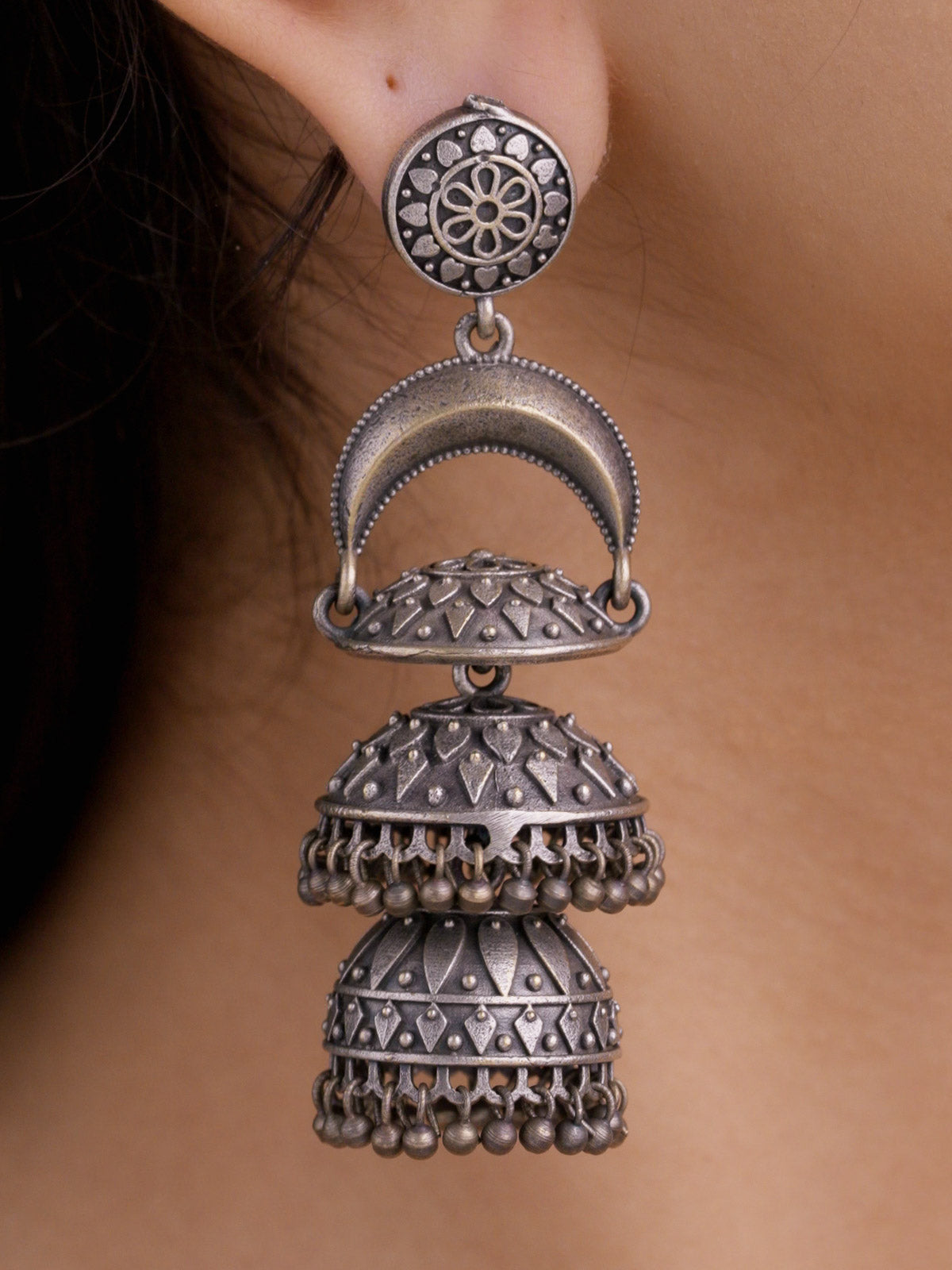 Close-up of a model wearing an oxidised tiered jhumka earring with a round floral stud, crescent arch connector, patterned double jhumka domes, and small round metal drops.
