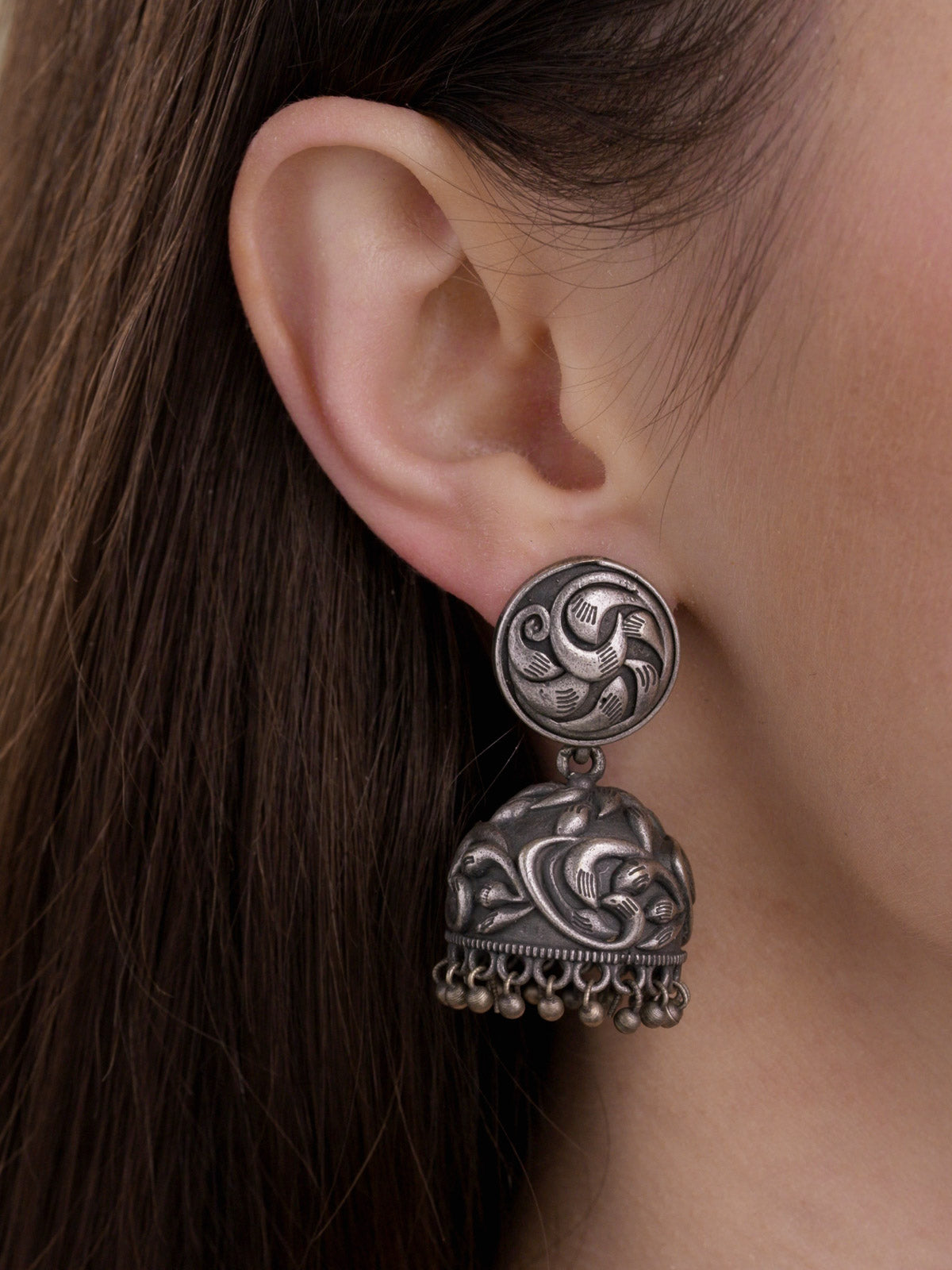 Model wearing an oxidised jhumka earring with a round carved stud, raised dome detailing and lower metal drops, close-up side view.