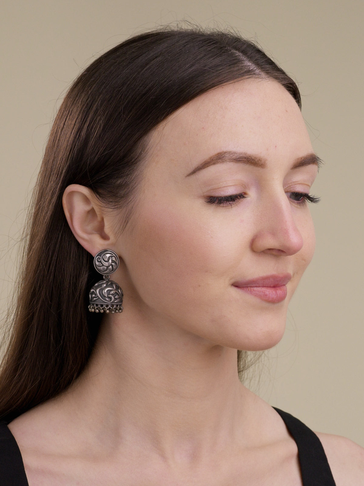 Model wearing an oxidised jhumka earring with a round carved stud, raised dome detailing and lower metal drops, side portrait view.