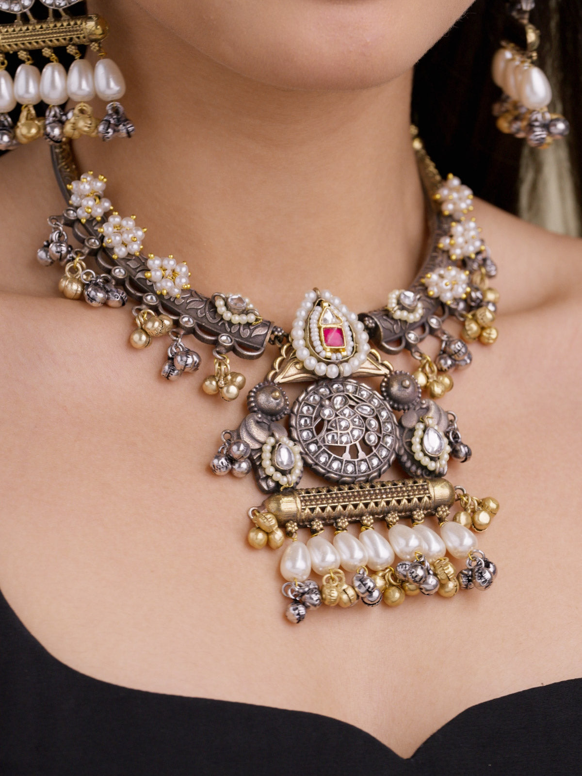 Close-up of a model wearing a multi tone oxidised necklace set with a round centrepiece, Ruby Red accent stone, pearl drops, gold and oxidised bell drops, floral pearl clusters, and matching earrings partly visible.