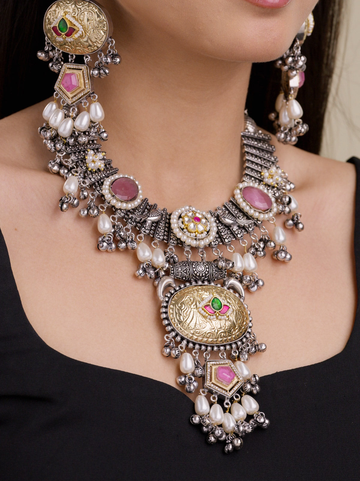 Model wearing a statement multi tone oxidised necklace set with antique gold oval medallions, pink stones, small green and ruby red accents, pearl drops, metal bead drops, and matching earrings in a close-up shot.