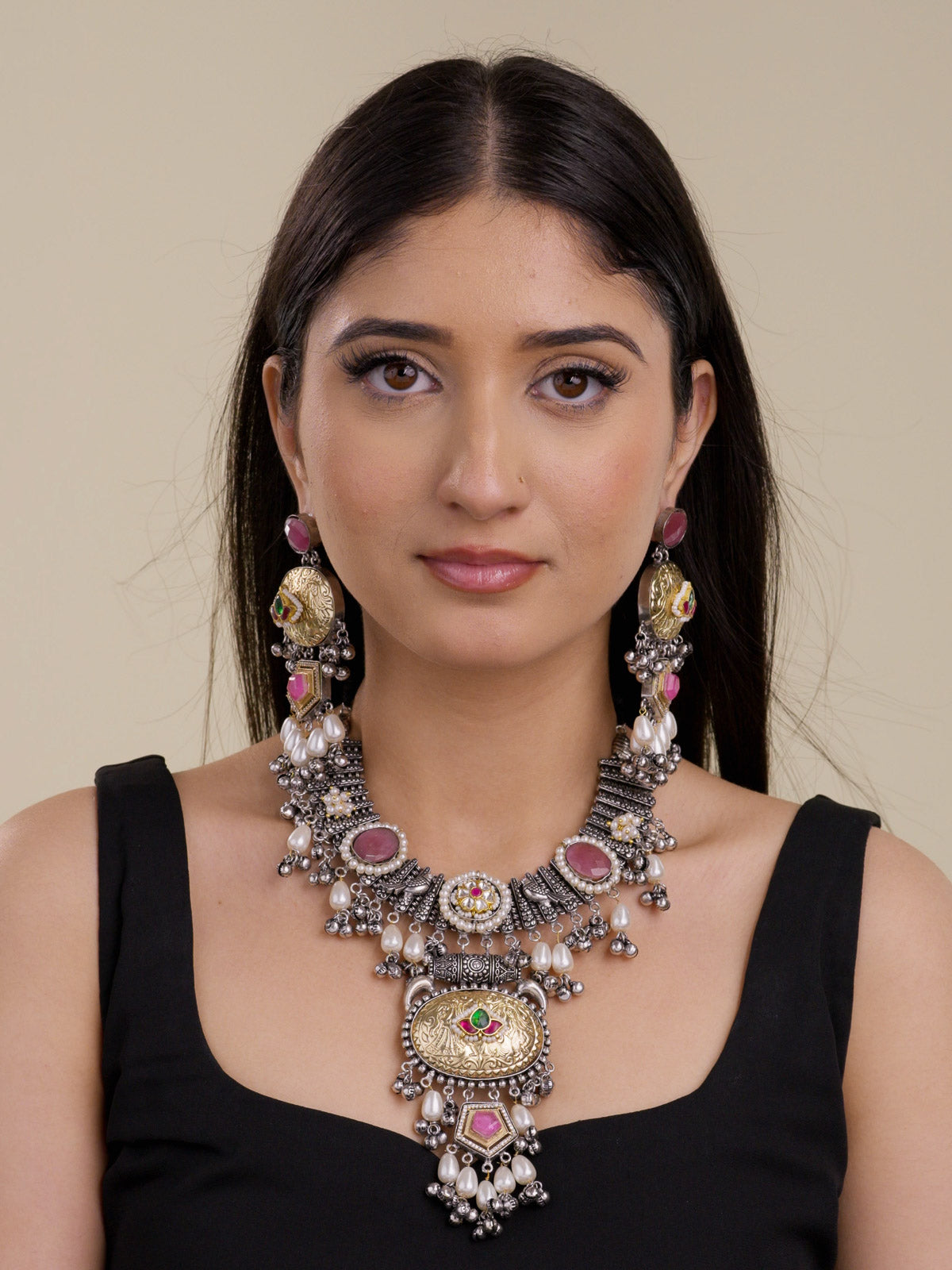 Model wearing a statement multi tone oxidised necklace set with antique gold oval medallions, pink stones, small green and ruby red accents, pearl drops, metal bead drops, and matching long earrings in a front portrait shot.